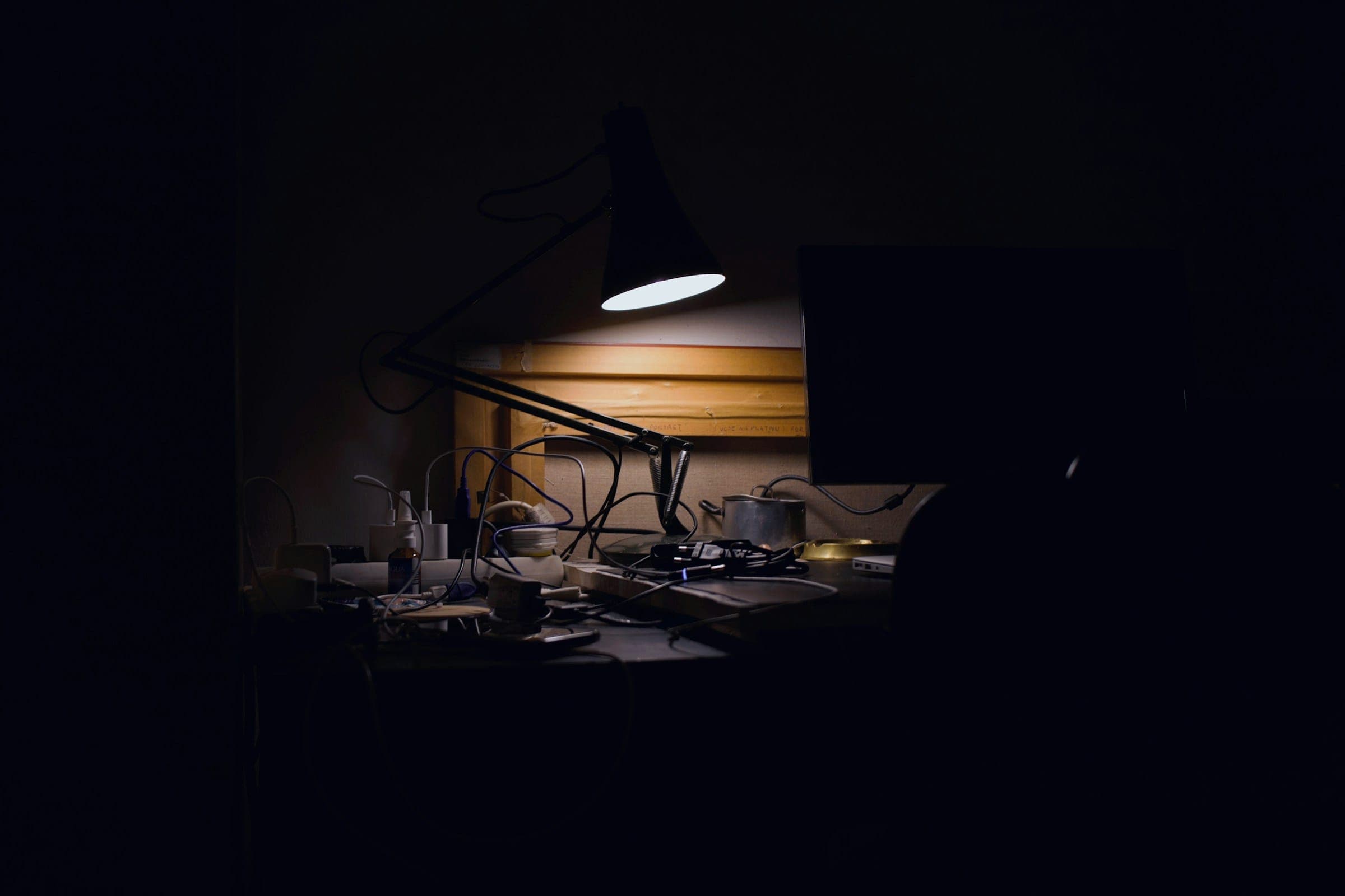 A dark room where a single desk lamp lights a cluttered workbench of cables and small tools.