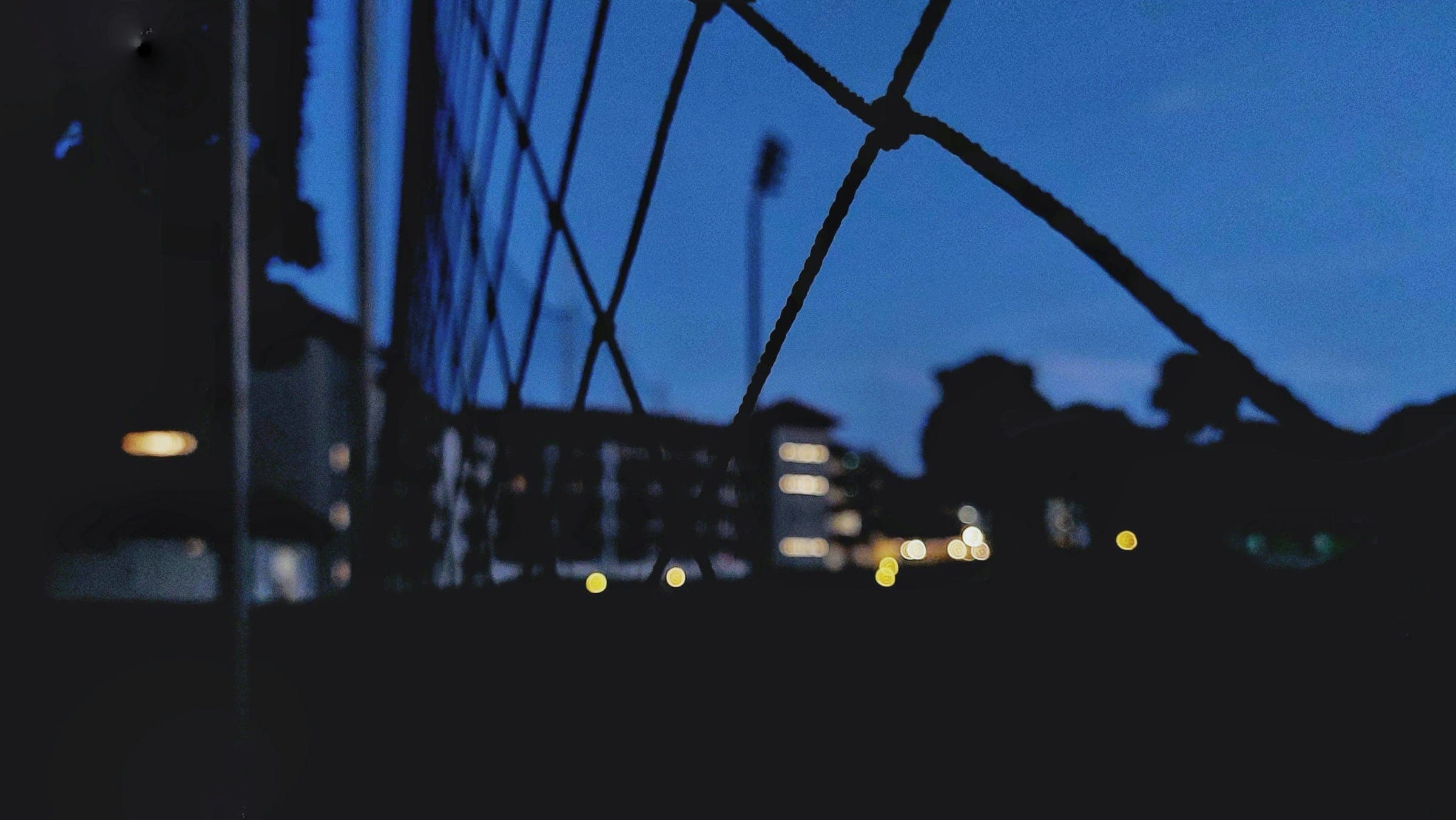 A city at dusk seen through a wire fence, buildings soft and out of focus.