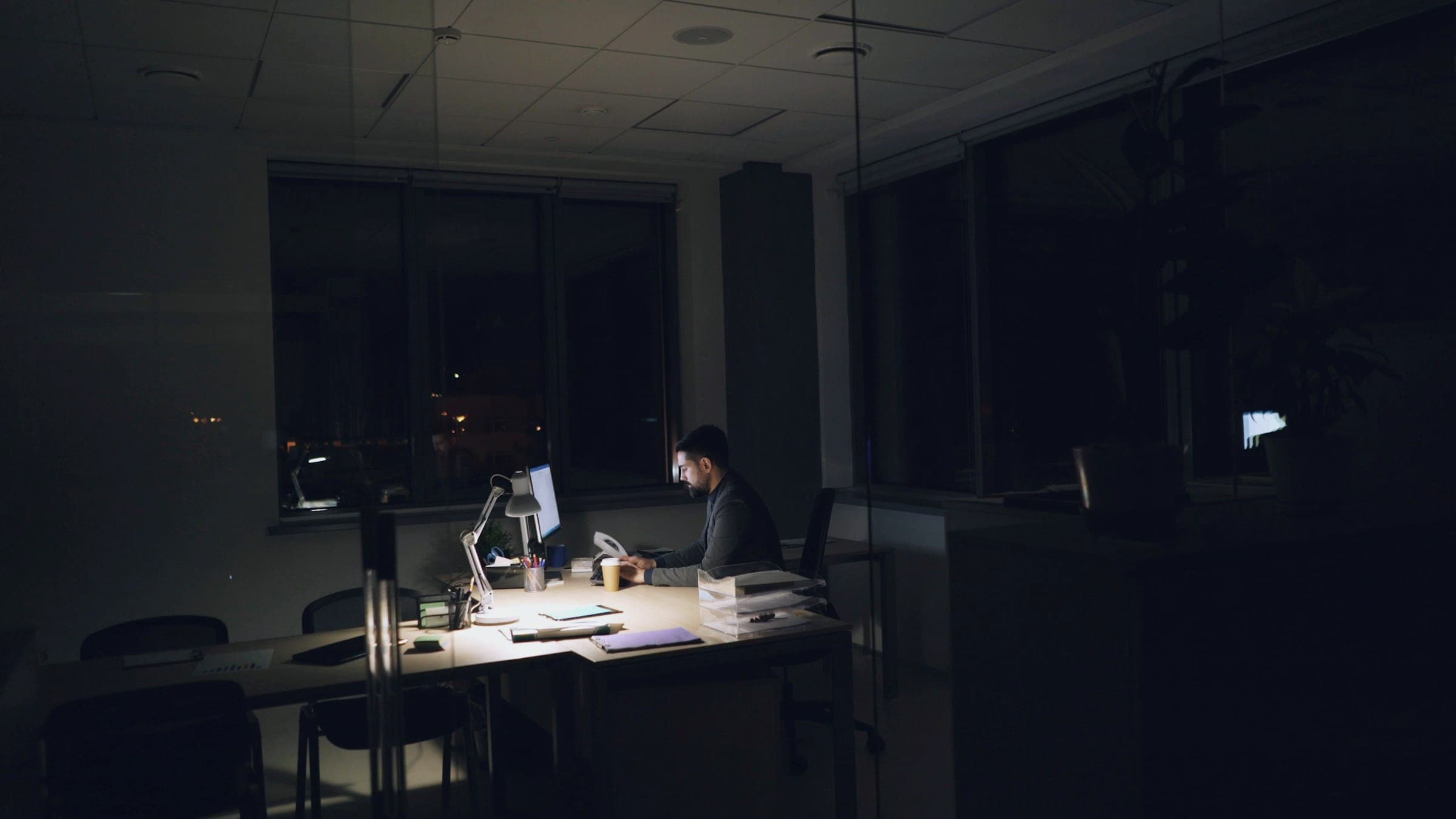 A lone worker at a desk lamp in an otherwise dark open-plan office at night.