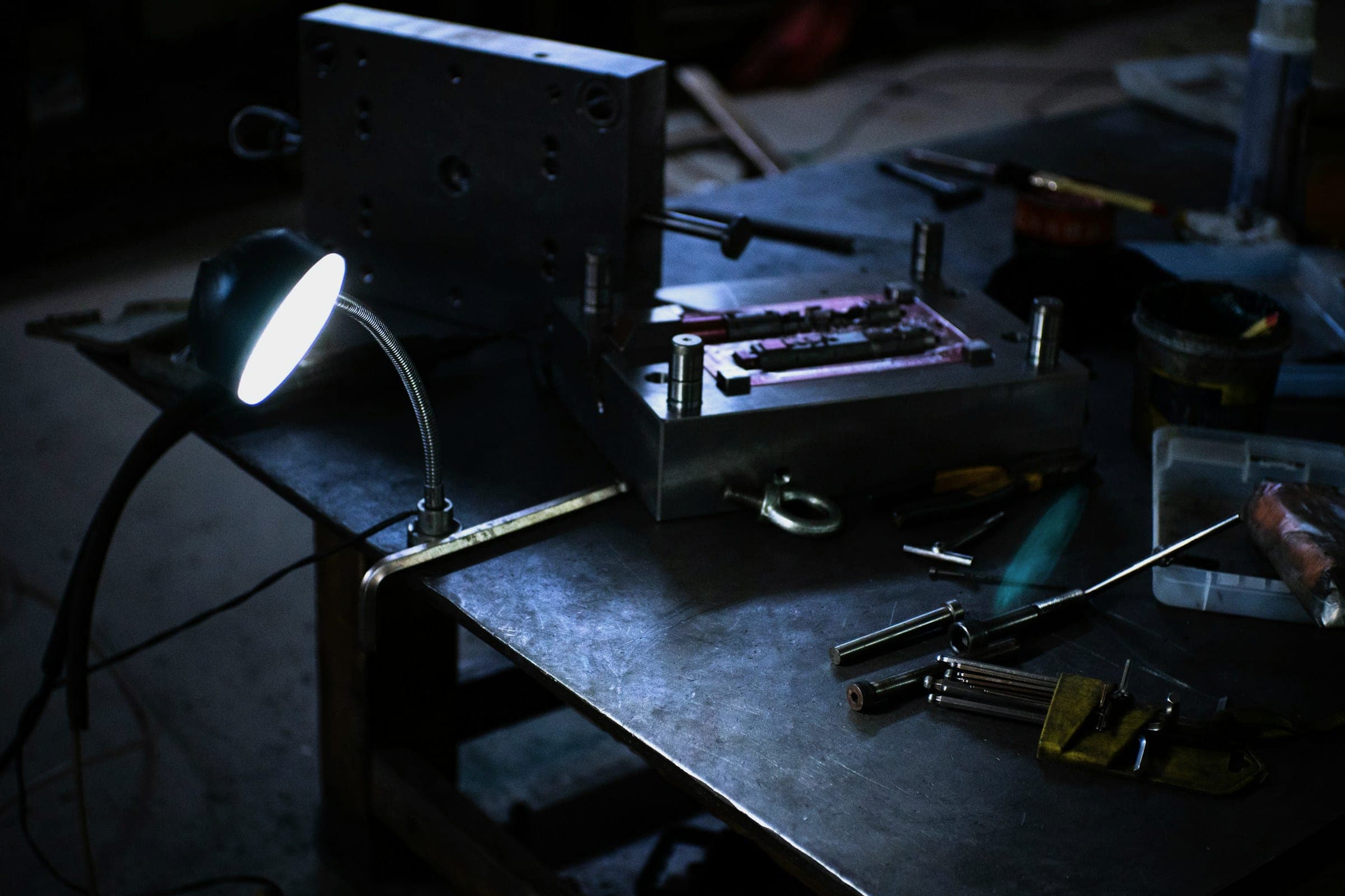 A workshop bench at night with tools, a machined part and a cool-white gooseneck lamp.